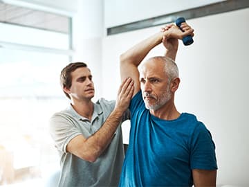A man in a blue shirt lifts a dumbbell overhead while another man assists, in a physical therapy session.