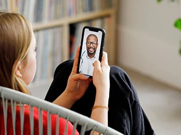 Person sitting on a chair, holding a smartphone with a video call on the screen, showing a smiling man wearing glasses and a stethoscope.