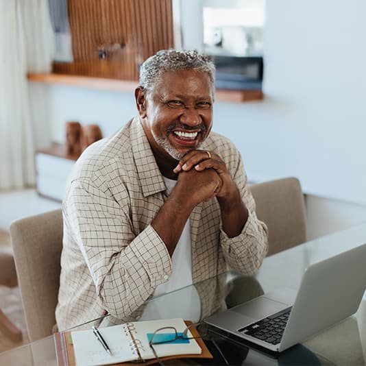 Smiling older man sitting at a glass table with a laptop and notebook, hands clasped under his chin, in a modern, bright interior.