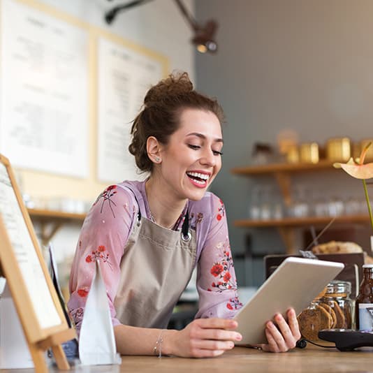 Smiling woman in a floral blouse and apron holds a tablet behind a cafe counter with menus and jars in the background.