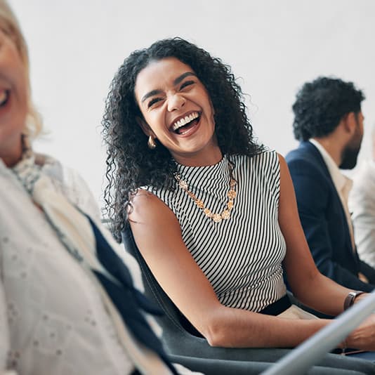 Smiling woman with curly hair in a striped top, sitting and laughing among a group of people.