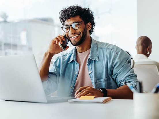 Smiling young man with glasses on phone while working at a laptop in a bright modern office.