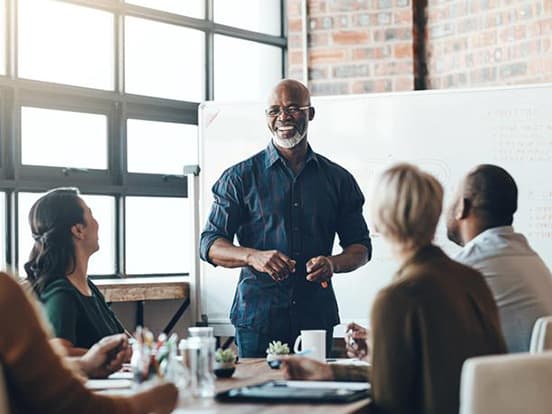 Man leading a meeting, standing by a whiteboard, engaging with four seated colleagues in a modern office setting.