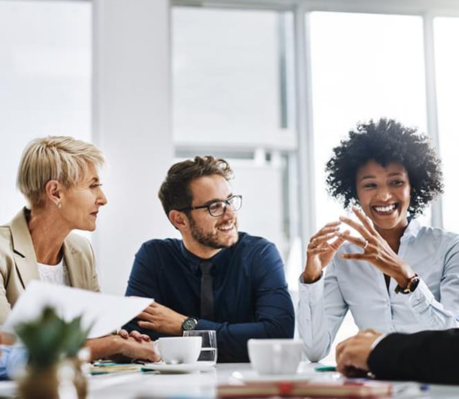 Three colleagues smiling and discussing around a conference table with coffee cups and documents.