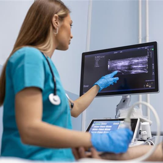 A healthcare professional in teal scrubs and blue gloves points at an ultrasound image on a monitor, holding a probe in a medical examination room.