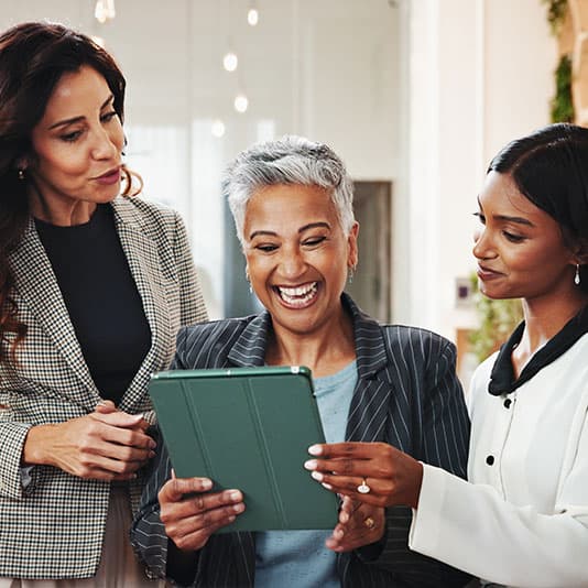 Three women smiling and looking at a tablet together in an office setting.