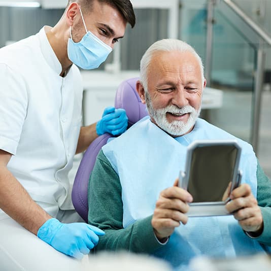 Older man smiling while holding a mirror, sitting in a dental chair. A dentist stands beside him, both appear pleased.