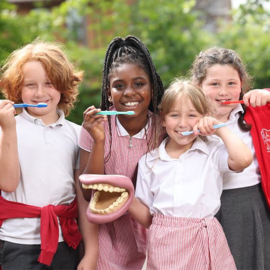 Four children smiling, holding toothbrushes, and a dental model outdoors. They wear school uniforms and stand in front of greenery.