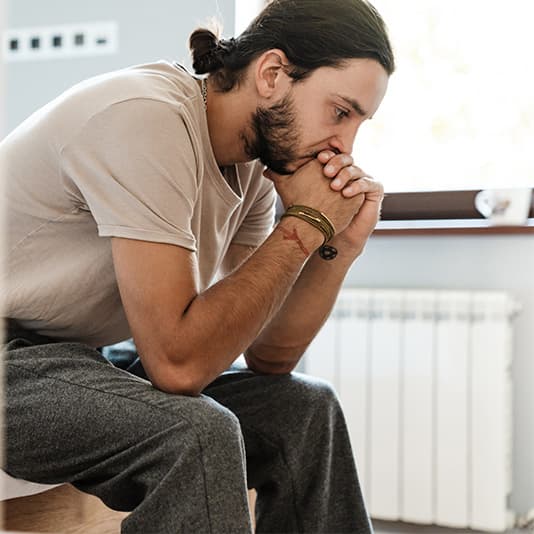 A man sitting on a bed, leaning forward with his hands clasped in front of his face, appearing deep in thought.