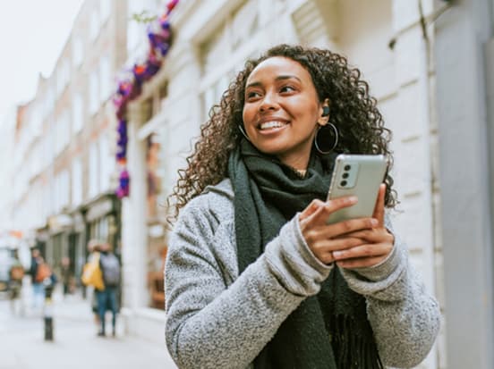 A smiling woman with curly hair holds a smartphone while walking on a city street. She wears a scarf and earbuds, with shops in the background.