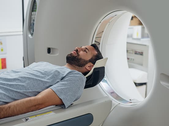 A man in a gray shirt lies on a table, entering a CT scanner in a medical facility.