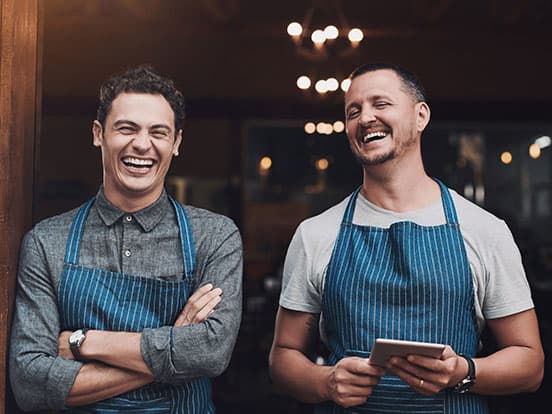 Two men in blue-striped aprons laughing in a café doorway, one with arms crossed and the other holding a tablet.