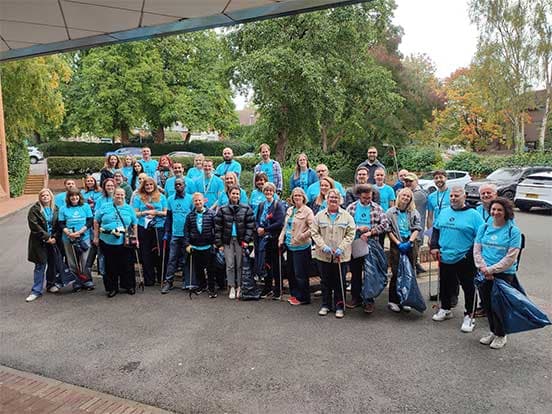 A group of people in matching blue shirts pose outdoors, holding litter-picking tools and bags, with trees and parked cars in the background.