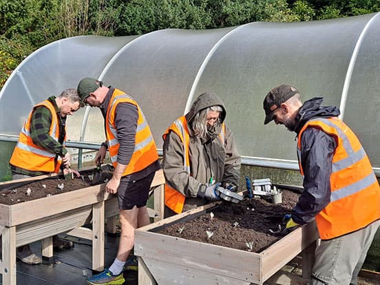 Four people in high-visibility vests plant seeds in raised garden beds under a greenhouse canopy.