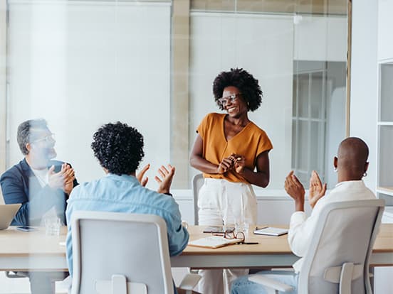 A woman stands smiling, receiving applause from three colleagues seated around a conference table in a modern office setting.