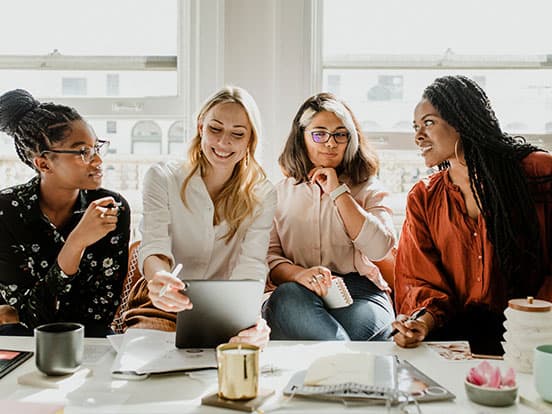 Four women are sitting together discussing around a table with notebooks and coffee cups, bathed in natural light.