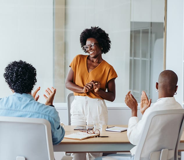 A person stands smiling at the head of a conference table while two seated individuals applaud in a modern office setting.
