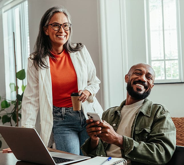 Smiling woman standing with coffee beside laughing man seated with phone and laptop in a bright office.